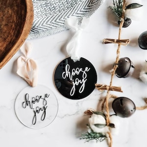 May include: Two round ornaments with the text "choose joy" written in black calligraphy. One ornament is clear acrylic and the other is black acrylic.