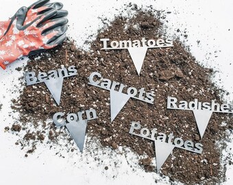 May include: A set of metal garden markers with the words "Tomatoes", "Beans", "Carrots", "Corn", "Radishes", and "Potatoes" printed on them. The markers are arranged on a bed of brown soil.