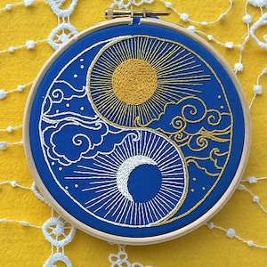 K&ouml;nnte beinhalten: Ein runder h&ouml;lzerner Stickrahmen mit blauem Stoff und einem handgestickten Yin-und-Yang-Motiv. Der obere Teil zeigt eine gelbe Sonne mit strahlenden Strahlen und goldenen Wolken, w&auml;hrend der untere Teil eine wei&szlig;e Mondsichel darstellt.