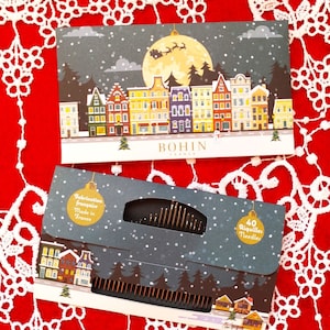 May include: Two rectangular paper packages of Bohin sewing needles featuring a festive winter town illustration with a full moon, snow, and a silhouette of Santa's sleigh. The packaging displays the text Bohin France and 40 Aiguilles Needles.