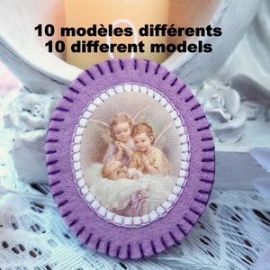 May include: Purple felt oval ornament with a white stitched border. The ornament features a picture of three angels with a baby. The text "10 modèles différents 10 different models" is printed above the ornament.