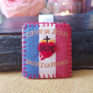 Pode incluir: Artigo de tecido bordado com um design de bandeira francesa. O artigo apresenta o texto "COEUR DE JESUS" e "SAUVEZ LA FRANCE" em fio dourado, juntamente com um coração vermelho e um emblema de cruz. Uma fita branca está presa no topo.