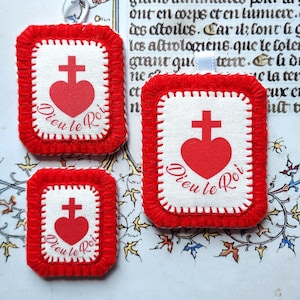 Puede incluir: Tres colgantes religiosos rojos y blancos. Cada colgante presenta un corazón rojo con una cruz y el texto francés "Dieu le Roi". Los colgantes tienen un borde rojo y costuras blancas.