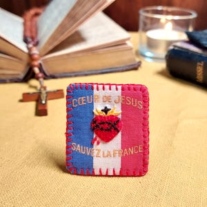 May include: A rectangular fabric patch featuring the French tricolour flag of blue, white, and red. It displays an embroidered red Sacred Heart with a cross and flames, with the gold text CŒUR DE JESUS SAUVEZ LA FRANCE stitched on the front.