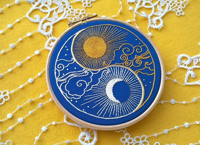 K&ouml;nnte beinhalten: Ein runder Holzstickrahmen mit einem blauen Stoffeinsatz und handgefertigter Stickerei. Das Motiv zeigt eine goldene Sonne und einen wei&szlig;en Halbmond in Yin-Yang-Anordnung, umgeben von goldenen und wei&szlig;en, wirbelnden Wolkenmustern.
