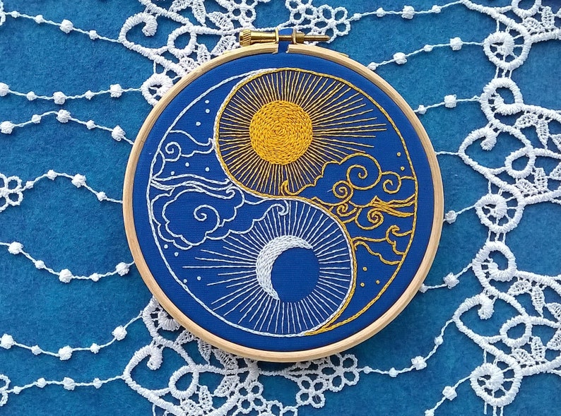 K&ouml;nnte beinhalten: Ein runder Stickrahmen aus Holz mit einem blauen Stoffeinsatz, der ein handgesticktes Yin-und-Yang-Motiv zeigt. Der obere Bereich enth&auml;lt eine goldgelbe Sonne mit strahlenden Strahlen und stilisierten Wolken, w&auml;hrend der untere Bereich eine wei&szlig;e Mondsichel mit Strahlen und goldenen Wolken darstellt.