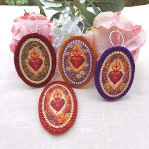 Puede incluir: Cuatro adornos ovalados con un diseño de Sagrado Corazón. Cada adorno presenta un corazón rojo con una llama, una cruz y una corona de espinas. Los adornos son de color rojo, naranja y morado, con lazos blancos para colgar.