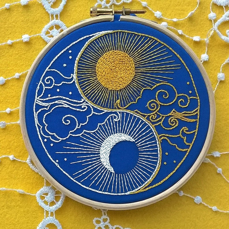 K&ouml;nnte beinhalten: Ein runder h&ouml;lzerner Stickrahmen mit blauem Stoff und einem handgestickten Yin-und-Yang-Motiv. Der obere Teil zeigt eine gelbe Sonne mit strahlenden Strahlen und goldenen Wolken, w&auml;hrend der untere Teil eine wei&szlig;e Mondsichel darstellt.
