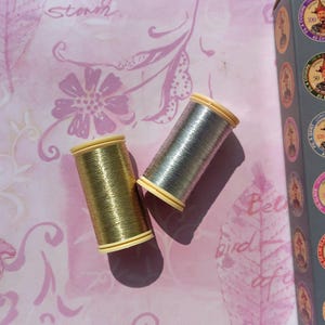 Pode incluir: Dois carretéis cilíndricos de fio metálico, um com acabamento dourado brilhante e outro com acabamento prateado cintilante, cada um com tampas de plástico de cor clara, posicionados na diagonal sobre uma superfície rosa com padrão floral.