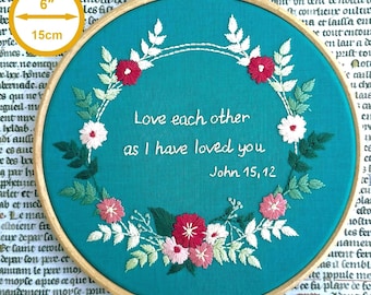 May include: A 6-inch circular wooden embroidery hoop containing teal fabric with a floral wreath stitched in shades of pink, white, and green. The center features white embroidered text reading: Love each other as I have loved you John 15,12.