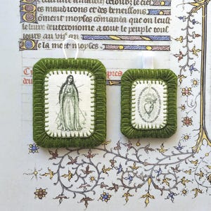May include: Two rectangular felt ornaments with olive green stitched borders. The larger ornament features a black and white illustration of a standing figure. The smaller ornament displays a heart illustration with the text Immaculate Heart of Mary.