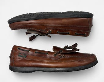 May include: A pair of brown leather boat shoes featuring a moccasin-style toe, dark brown laces threaded through metal eyelets, and a black rubber sole. A small green tag with the text Sebago is visible on the side of the shoe.