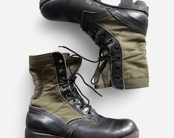 May include: A pair of black and olive green combat boots featuring a leather lower construction, canvas side panels, black laces, metal eyelets, and thick, lugged rubber soles. The boots have a high-top design with reinforced stitching.