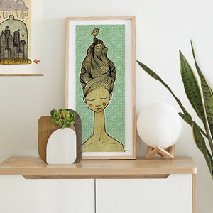 Op de afbeelding: Een ingelijste illustratie van een vrouw met een vogel die op haar uitgebreide kapsel zit. De achtergrond is lichtgroen met een herhalend patroon. De vrouw heeft haar ogen gesloten en een lange nek. De illustratie staat op een houten plank met een kleine vetplant in een witte vaas en een witte maanlamp op een houten standaard.