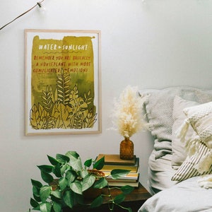 Pode incluir: Uma impressão emoldurada com fundo amarelo e texto preto que diz "Water + Sunlight. Remember you are basically a houseplant with more complicated emotions." A impressão apresenta um desenho de folhas e plantas verdes.