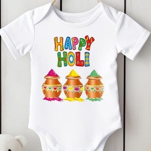 Könnte beinhalten: Weißer Baby-Strampler mit einem bunten "Happy Holi"-Motiv und drei Tonkrügen, die mit buntem Pulver gefüllt sind.