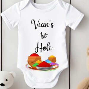 Könnte beinhalten: Weißer Baby-Strampler mit schwarzem Text "Vian's 1st Holi" und einer bunten Illustration von Holi-Pulver und Schalen.
