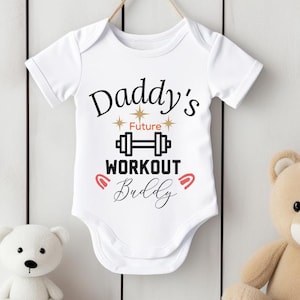 Peut inclure: Combinaison blanche pour bébé avec un texte noir qui dit "Daddy's Future Workout Buddy" avec un graphique d'haltère et des étoiles dorées. La combinaison est suspendue à un cintre en bois.