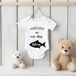May include: White baby onesie with the text "DADDY LOVES me more than fishing" and a black fish graphic. A white teddy bear and a tan teddy bear are on either side of the onesie.