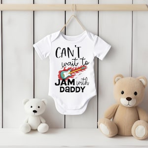 May include: White baby bodysuit featuring the text "CAN'T wait to JAM with DADDY" and a guitar graphic. A white teddy bear and a tan teddy bear are on either side of the bodysuit.