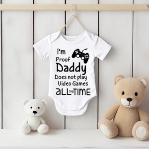 Puede incluir: Un body blanco para bebé con texto negro que dice "Soy la prueba de que papá no juega a los videojuegos TODO EL TIEMPO" y un gráfico de un mando de videojuegos negro.