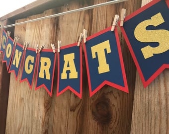 May include: A banner spelling "CONGRATS" is strung across a wooden background. Each pennant is navy blue with a red border and gold glitter letters. The banner is held up by small wooden clothespins.