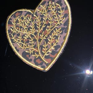 May include: Heart-shaped embroidered patch with a gold border. The patch features a leopard print background and a gold embroidered tree design. The patch is likely for clothing or accessories.