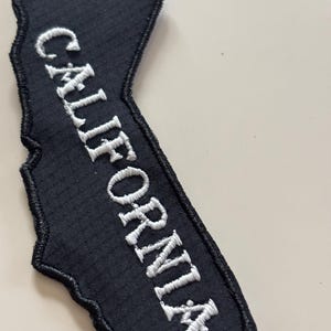 Puede incluir: Parche negro con la forma de California con texto bordado blanco "CALIFORNIA". El parche tiene un borde negro y una superficie texturizada.