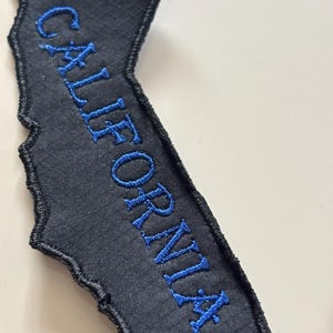 Puede incluir: Un parche negro con la forma de California con texto bordado en azul que dice "CALIFORNIA". El parche tiene una superficie texturizada y un borde cosido.