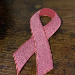 May include: A pink ribbon patch with a stitched design. The ribbon is a symbol of breast cancer awareness. The patch is made of fabric and has a detailed, textured appearance.