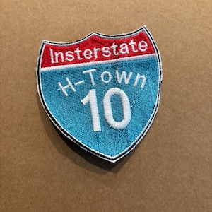 Puede incluir: Parche bordado en forma de escudo, con el texto "Interstate" en rojo, "H-Town" y el número "10" en blanco sobre un fondo azul. El parche tiene un borde negro.