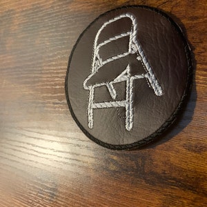 Puede incluir: Un parche redondo negro con un contorno bordado en blanco de una silla. El parche tiene un borde negro y está sobre un fondo de madera. El diseño de la silla es simple y estilizado.