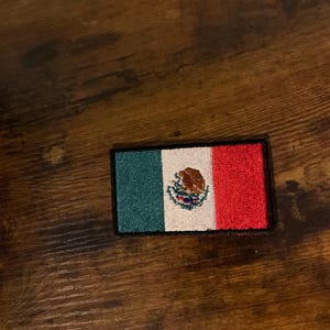 Puede incluir: Parche bordado con la bandera mexicana. El parche tiene forma rectangular con franjas verticales verdes, blancas y rojas. La franja blanca central muestra el escudo de armas mexicano. El parche está bordeado en negro.