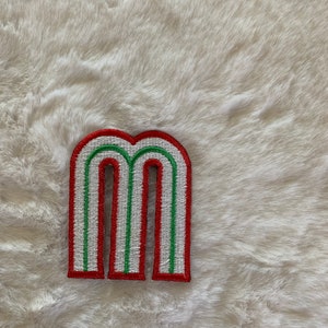 May include: Embroidered patch in the shape of the letter "M". The patch has a white background with red and green accents. The edges are outlined in red. The patch is on a white, fluffy surface.