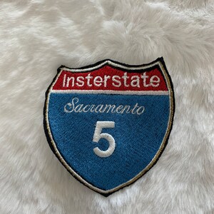 Puede incluir: Parche bordado con forma de señal de carretera. El parche presenta una pancarta roja con la palabra "Interstate" y una pancarta azul con "Sacramento 5". El parche tiene un borde negro y un contorno dorado.