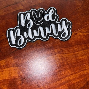 Pode incluir: Um patch bordado em preto e branco com o texto "Bad Bunny" em uma escrita estilizada. O design inclui um ícone de cabeça de coelho acima da palavra "Bad". O patch tem uma borda branca e está sobre uma superfície de madeira.