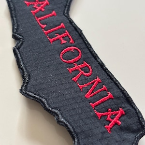 Puede incluir: Parche bordado negro con la forma de California. La palabra "CALIFORNIA" está cosida en rojo en el parche. El parche tiene una superficie texturizada y un borde negro.