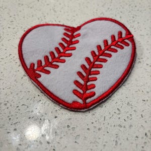 Op de afbeelding: Een hartvormige honkbalpatch met een witte kern en rode stiksels. De rode stiksels omlijnen het hart en bootsen de naden van een honkbal na. De patch is waarschijnlijk gemaakt van vilt of een vergelijkbare stof.
