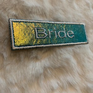 May include: A rectangular patch with the word "Bride" embroidered in white on a teal and gold iridescent background. The patch has a silver border and is set against a fluffy, light-colored surface.