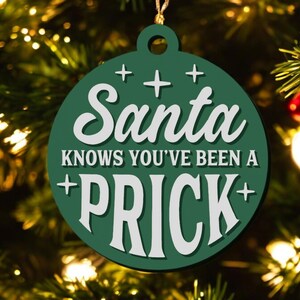 May include: Green Christmas ornament with the text "Santa Knows You've Been a Prick" in white. The ornament is round with a loop for hanging and is decorated with cross shapes. The background is a Christmas tree.