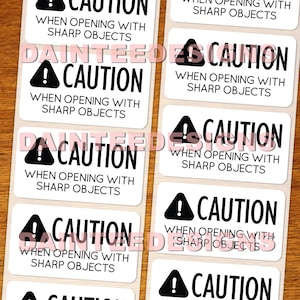 Puede incluir: Una hoja de pegatinas rectangulares blancas con texto negro y un símbolo de precaución negro. Cada pegatina dice "CAUTION" encima del texto "WHEN OPENING WITH SHARP OBJECTS". Las pegatinas están dispuestas sobre una superficie de madera.