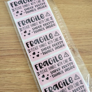 May include: Pink rectangular stickers with the word "FRAGILE" in black, along with the phrases "PLEASE HANDLE WITH CARE" and "SUPER PRETTY THINGS INSIDE." Each sticker has a warning symbol and heart graphics. The stickers are in a clear plastic sleeve.
