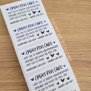 May include: A roll of white rectangular stickers with black text. Each sticker reads "Epoxy Pen Care" and provides care instructions. The stickers also include small black heart illustrations. The brand name "Daintee Designs" is visible.