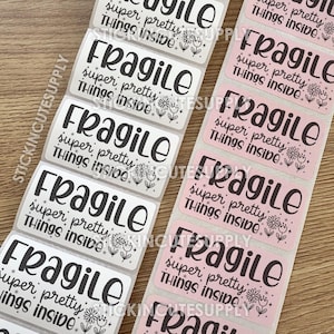 May include: Two rolls of rectangular stickers. One roll has white stickers with the word "Fragile" in black, along with the text "super pretty things inside." The other roll has pink stickers with the same text.
