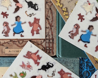 Op de afbeelding: Vellen stickers met illustraties van beren, een meisje in een blauwe jurk, een kat, een vos, een kip en een gans. De stickers liggen op vintage kinderboeken met gouden letters.