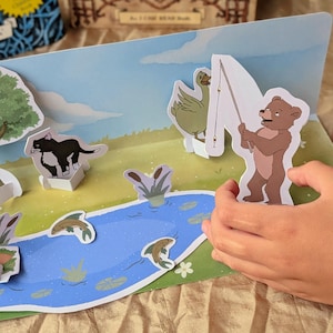 Può includere: Una scena di libro pop-up con un orso bruno che tiene una canna da pesca, un'anatra verde e un gatto bianco e nero. L'illustrazione mostra uno stagno blu con pesci che saltano, erba verde e un cielo azzurro su una base piatta.