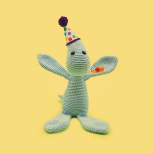 May include: A light blue crocheted kangaroo wearing a colorful party hat with a purple pom-pom. The kangaroo has black eyes and a small orange party horn in its mouth.