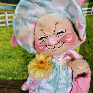 May include: A whimsical bunny doll with a painted face, wearing a floral print dress and hat. The doll holds a yellow flower and a pink purse. The bunny has rosy cheeks and a smiling expression, perfect for Easter decor.