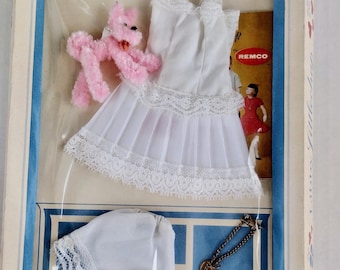 May include: A white sleeveless dress with lace trim and a pleated skirt, a pink pipe cleaner poodle, a white bonnet with lace edging, and a gold-tone chain bracelet, all packaged in a box with a Remco logo and a printed image of a child.