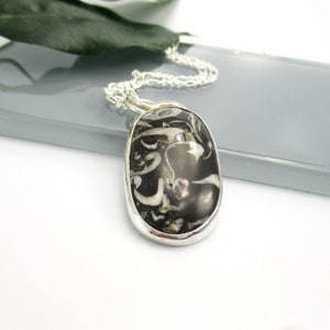 May include: A silver chain necklace with a black and white oval pendant. The pendant is made of a natural stone with a swirling pattern.